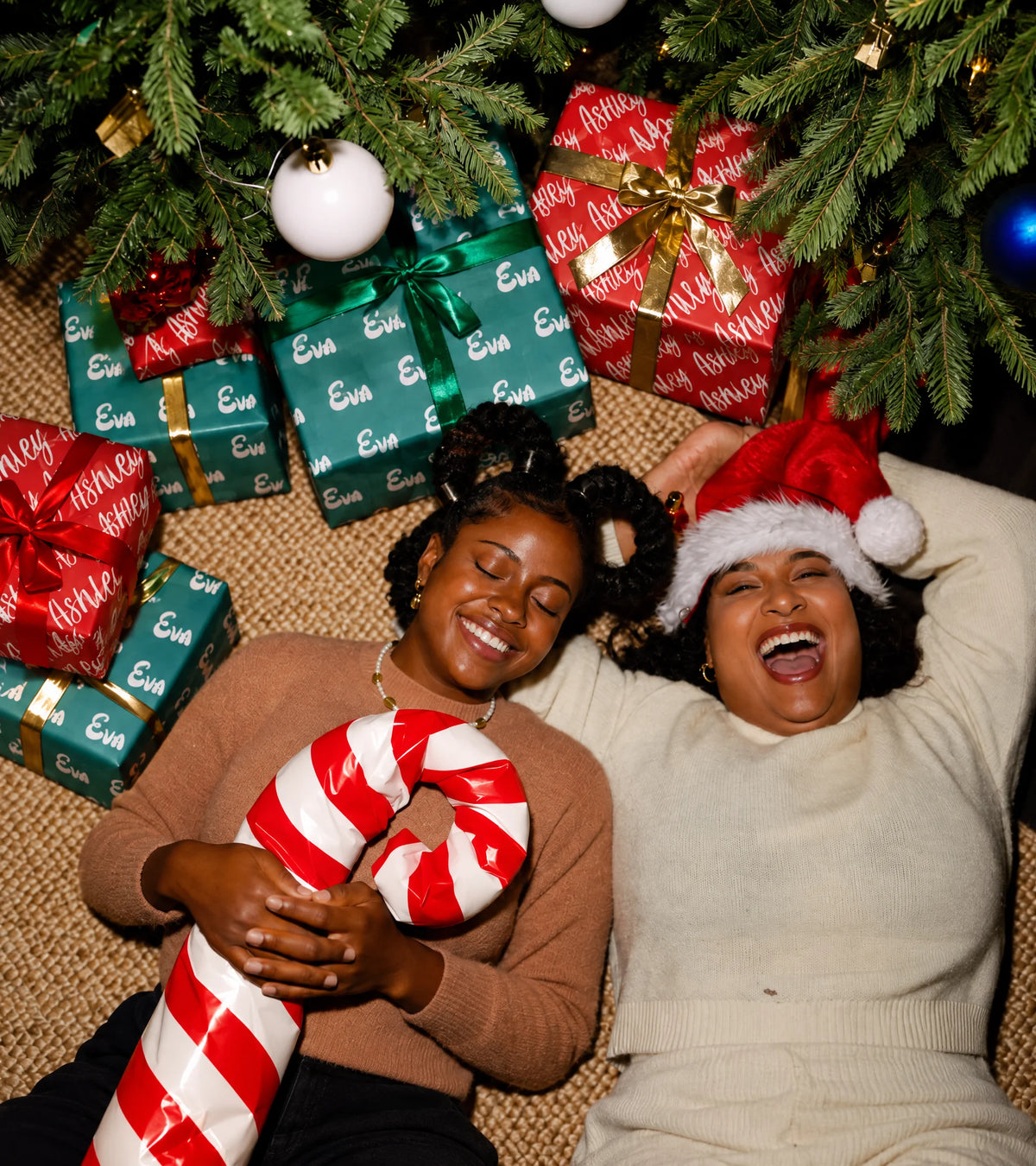 Two friends laughing under a Christmas tree beside gifts wrapped in personalized Paperbloom paper